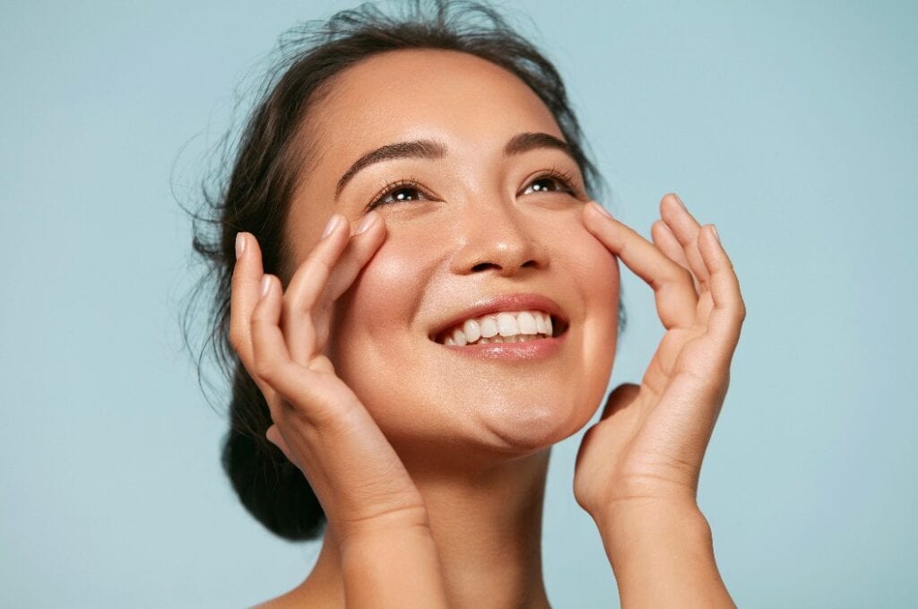 Skin care. Woman with beauty face touching healthy facial skin portrait. Beautiful smiling asian girl model with natural makeup touching glowing hydrated skin on blue background closeup